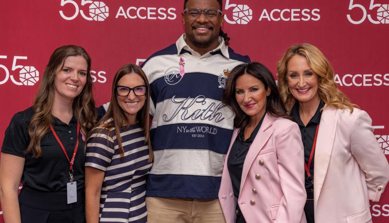 Five people smile and pose together in front of a red step-and-repeat backdrop featuring the ACCESS 55th anniversary logo. The group includes four women and one man, with the man standing in the center wearing a navy-and-white striped shirt.