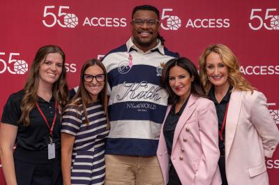 Five people smile and pose together in front of a red step-and-repeat backdrop featuring the ACCESS 55th anniversary logo. The group includes four women and one man, with the man standing in the center wearing a navy-and-white striped shirt.
