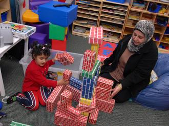 A young girl in a red outfit and a woman wearing a patterned hijab sit on the floor of a classroom, playing together with colorful foam building blocks designed to look like bricks. They are constructing a tall, elaborate structure surrounded by toys, bookshelves, and classroom supplies.